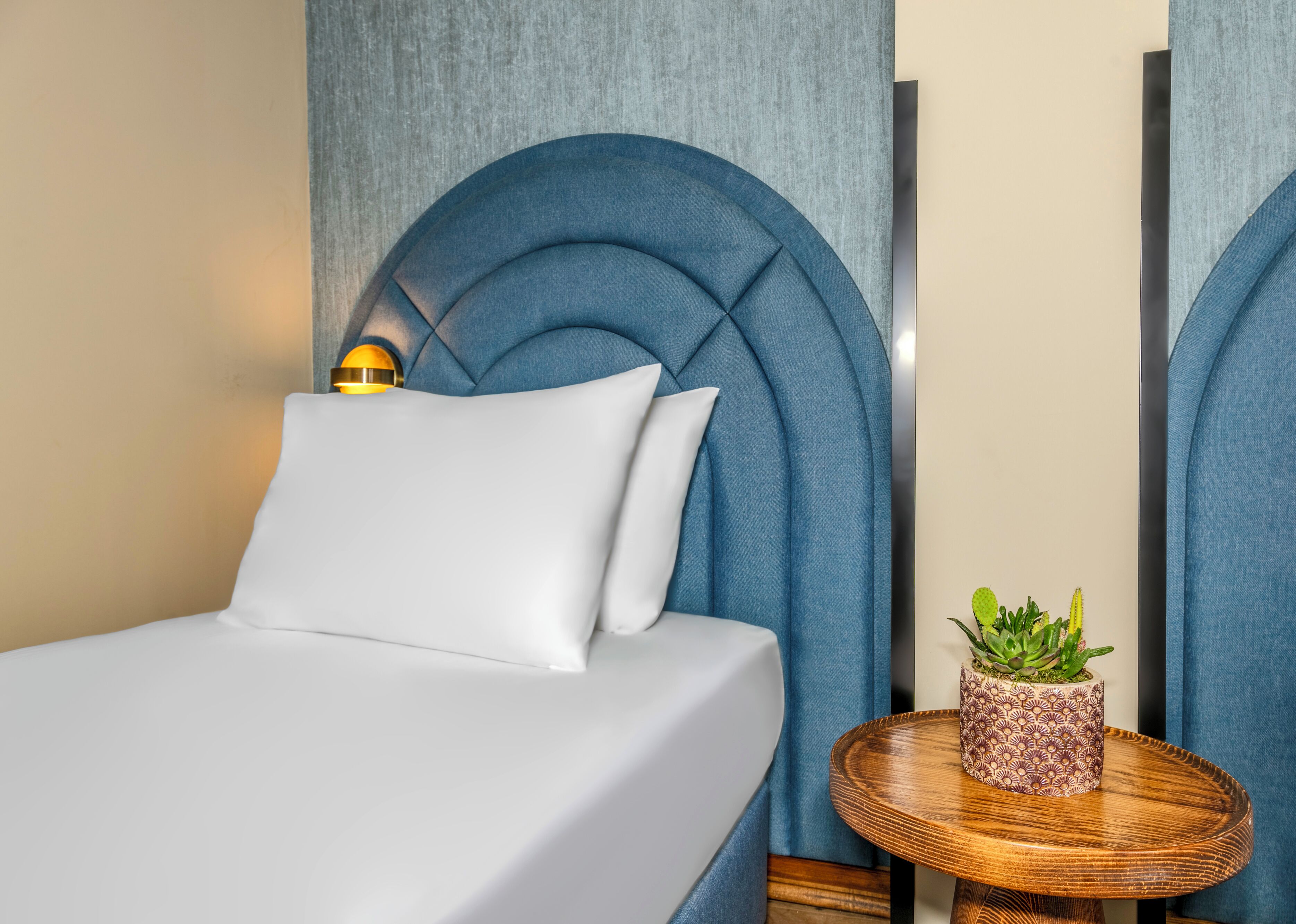 A neatly made bed with white linens and a blue headboard. There is a small round wooden table with a potted plant beside the bed.