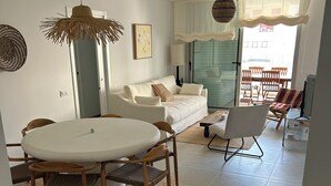 En stue med en hvid sofa, et rundt spisebord med stole, et fjernsyn og et stort vindue med gardiner.