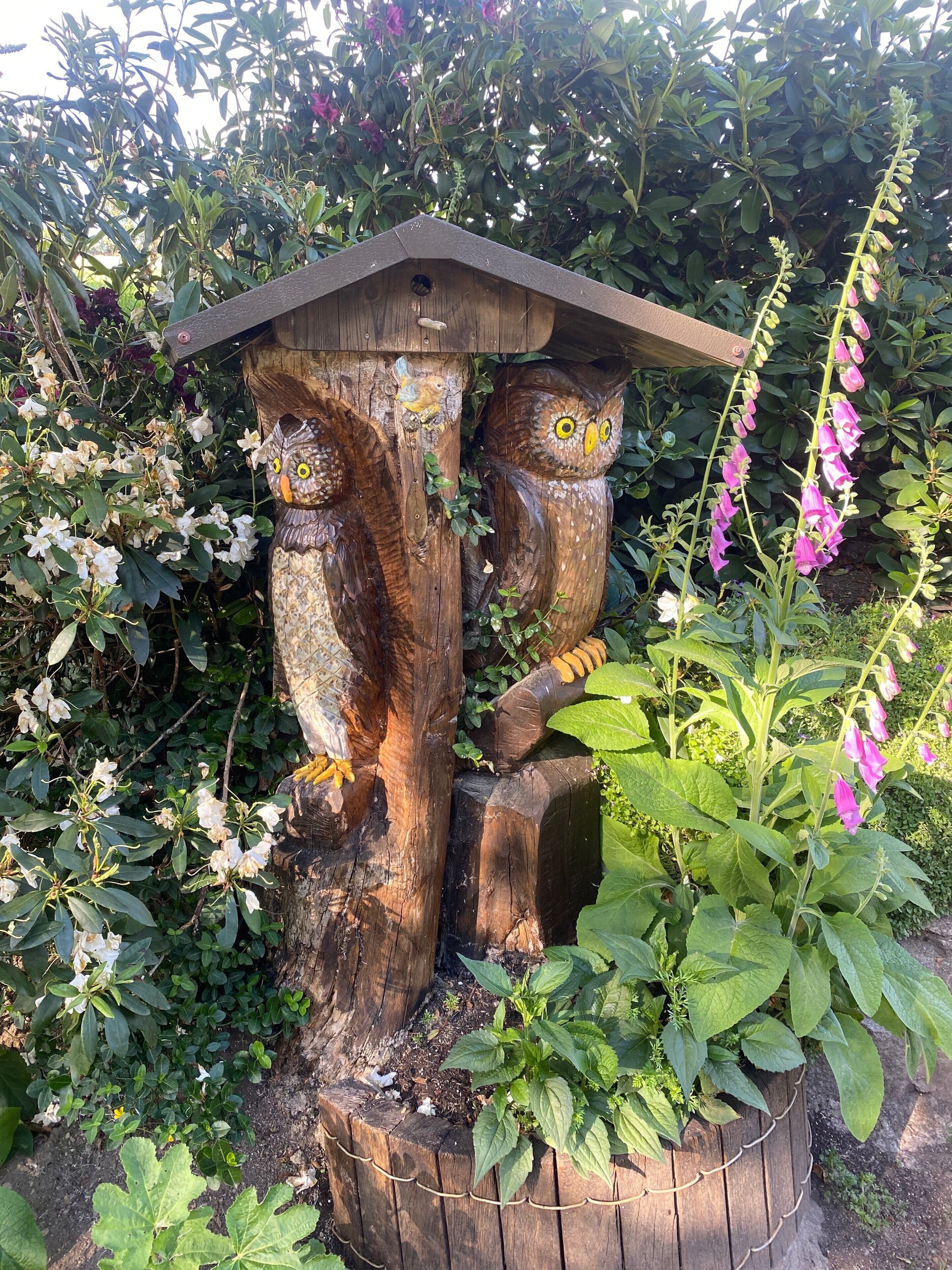 Una estructura de casa para pájaros de madera con techo, rodeada de follaje y plantas con flores.