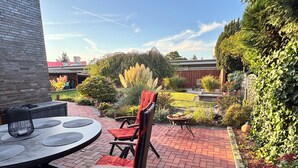 Une terrasse avec une table ronde et des chaises, entourée d’un chemin en briques et d’une végétation luxuriante.
