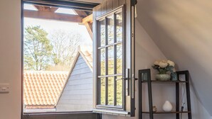 A room with a dark door, a window with a view of a tiled roof, a wicker basket, and a shelf with decorative items. - Peaceful Stylish Entire Apartment - Views, Gardens, Firepit, Balcony (Holt)