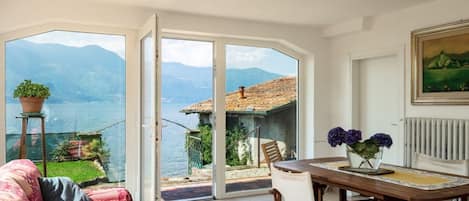 Ein Zimmer mit einer großen Schiebeglasdoor, die auf einen Balkon mit Blick auf einen See und Berge führt. Im Inneren befindet sich ein Holztisch mit Stühlen und ein Sofa.