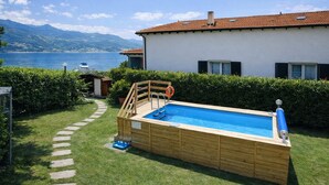 Ein oberirdischer Pool aus Holz mit einer Leiter, eine kleine Gartenhütte und im Hintergrund ein See und Berge.