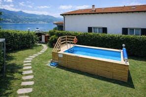 Ein oberirdischer Pool aus Holz mit einer Leiter, eine kleine Gartenhütte und im Hintergrund ein See und Berge.