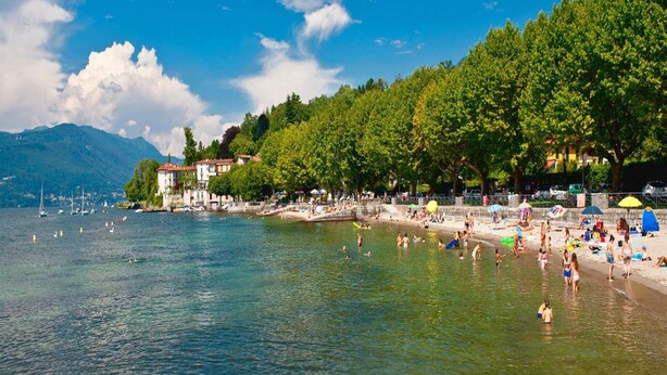 Ein Strand mit klarem Wasser, Menschen beim Schwimmen und Sonnenbaden, und Gebäuden entlang der Küste.