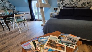 Un dormitorio con cama, una mesita de centro de madera con folletos de viajes, un escritorio con silla y una ventana con cortinas.