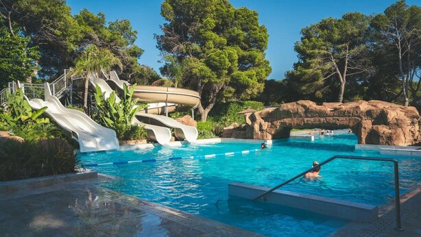 A swimming pool with slides and a boat, surrounded by trees and rocks.