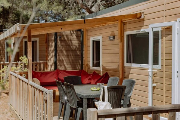 A wooden cabin with a covered patio area featuring a table and chairs, a red cushioned bench, and a small window.