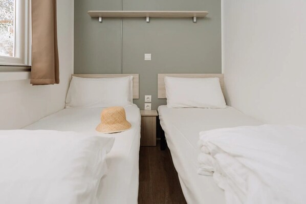 A twin room with two single beds, a small nightstand, a wall-mounted shelf, and a window with a curtain.