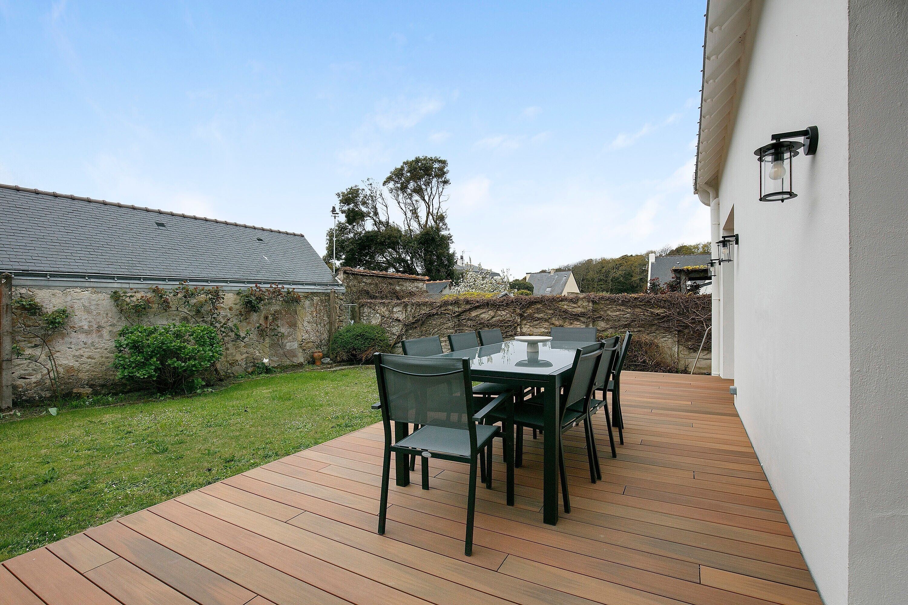 A wooden deck with a table and chairs, a white wall, and a stone wall in the background.