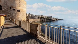 Um calçadão à beira-mar com uma torre de pedra, um deck de madeira e vista para edifícios do outro lado da água.