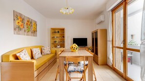 Uma sala de estar com sofá, mesa de jantar, cadeiras, televisão, estante de livros e uma janela grande.