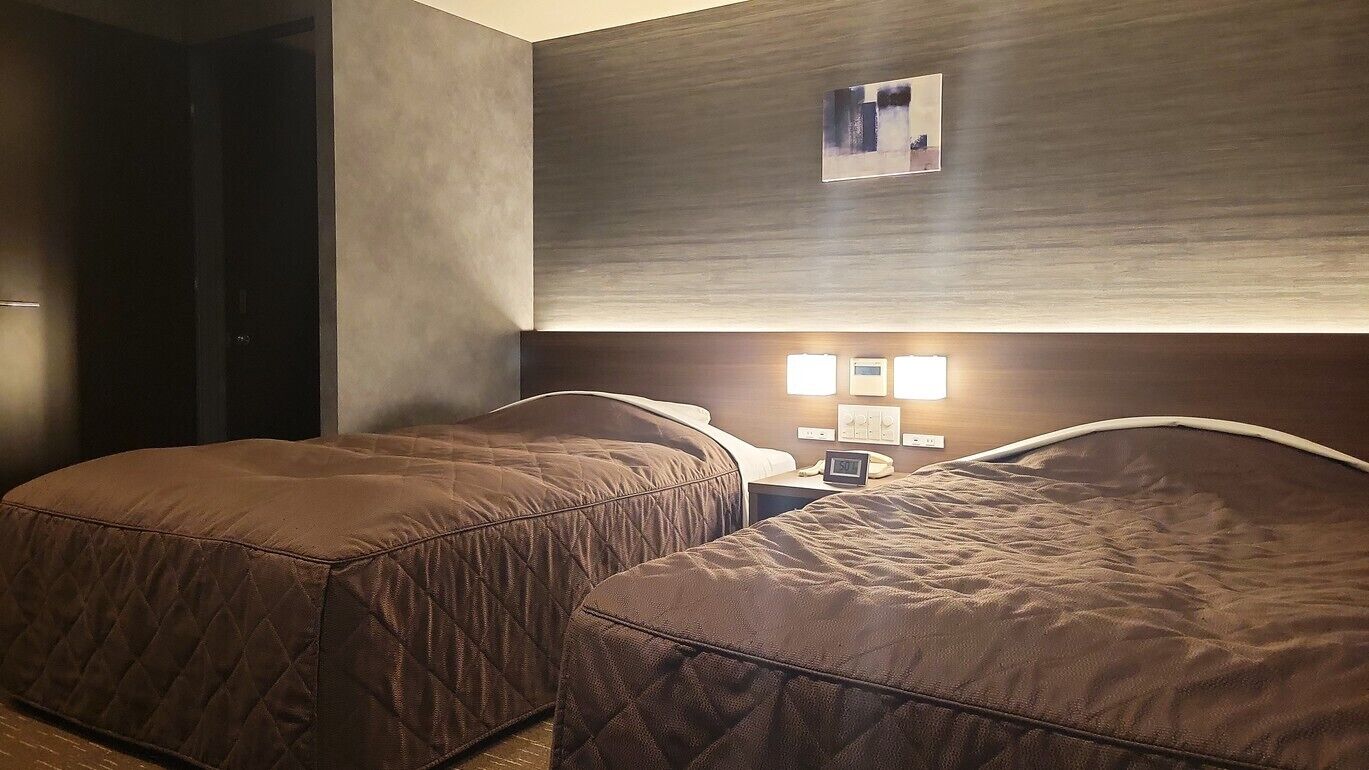 Quarto de hotel com duas camas, uma mesa de cabeceira com relógio e luminária fixada na parede.