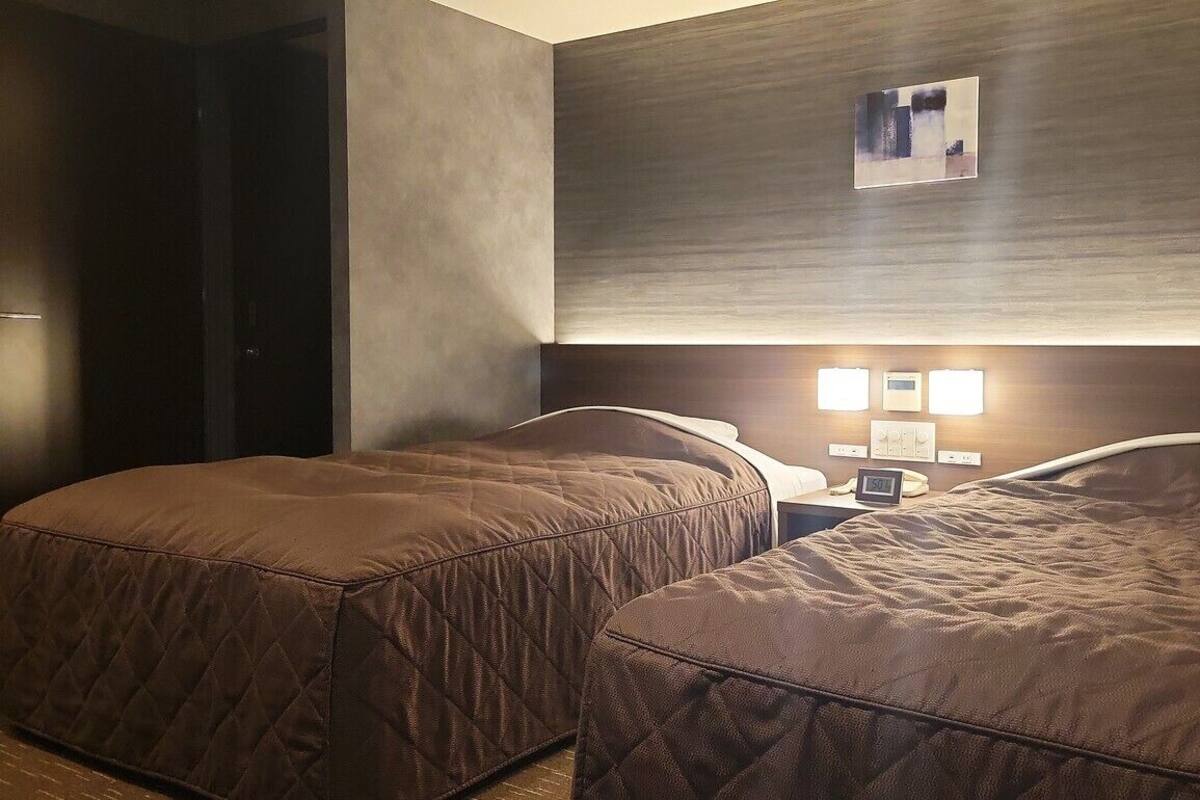 Una habitación de hotel con dos camas, una mesita de noche con un reloj y una lámpara fijada a la pared.