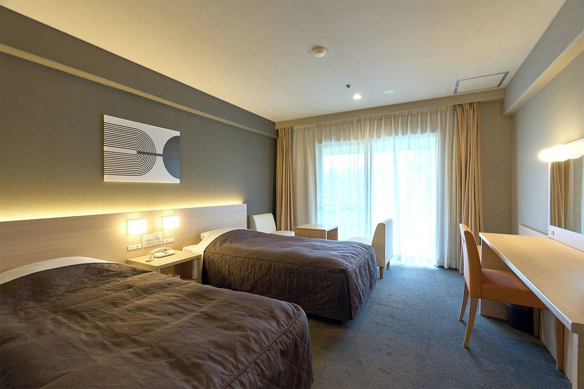 A hotel room with two beds, a desk, a chair, a lamp, and a large window with curtains.