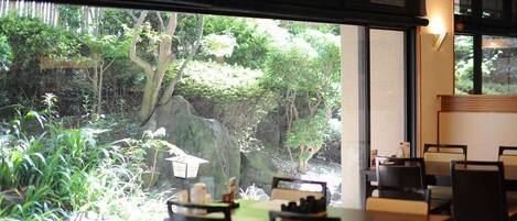 A restaurant with a view of a rocky garden and trees. Tables and chairs are arranged for dining.