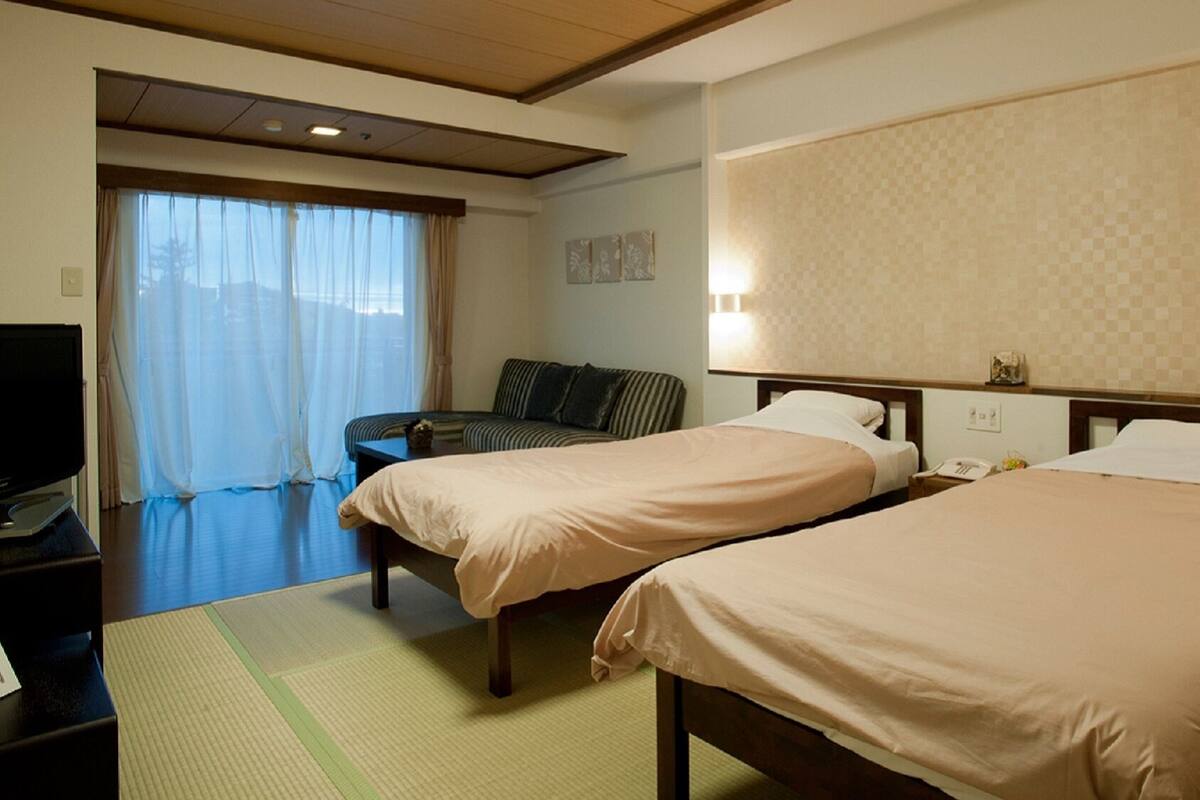 A hotel room with two beds, a television, a sofa, and a window with curtains.