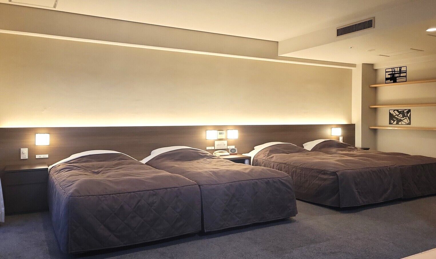 A hotel room with two beds, bedside lamps, a nightstand, a shelf with a picture, and a vent on the ceiling.