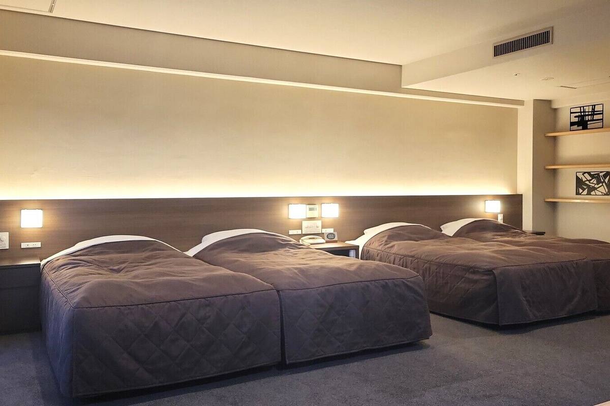 A hotel room with two beds, bedside lamps, a nightstand, a shelf with a picture, and a vent on the ceiling.