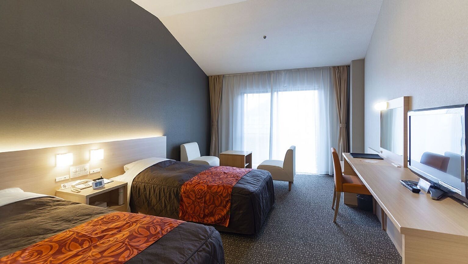 Quarto de hotel com duas camas, uma mesa com computador, uma cadeira, uma televisão e uma janela com cortinas.