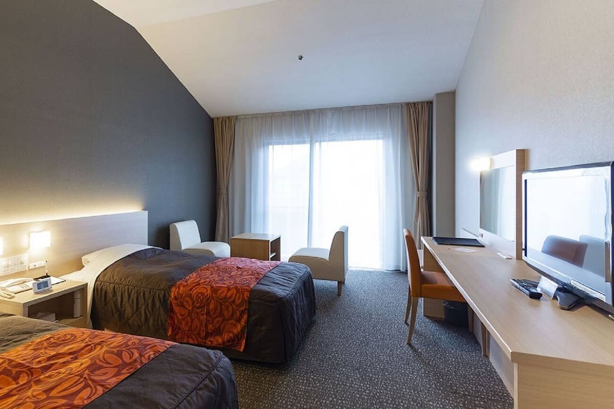 Habitación de hotel con dos camas, un escritorio con computadora, una silla, un televisor y una ventana con cortinas.