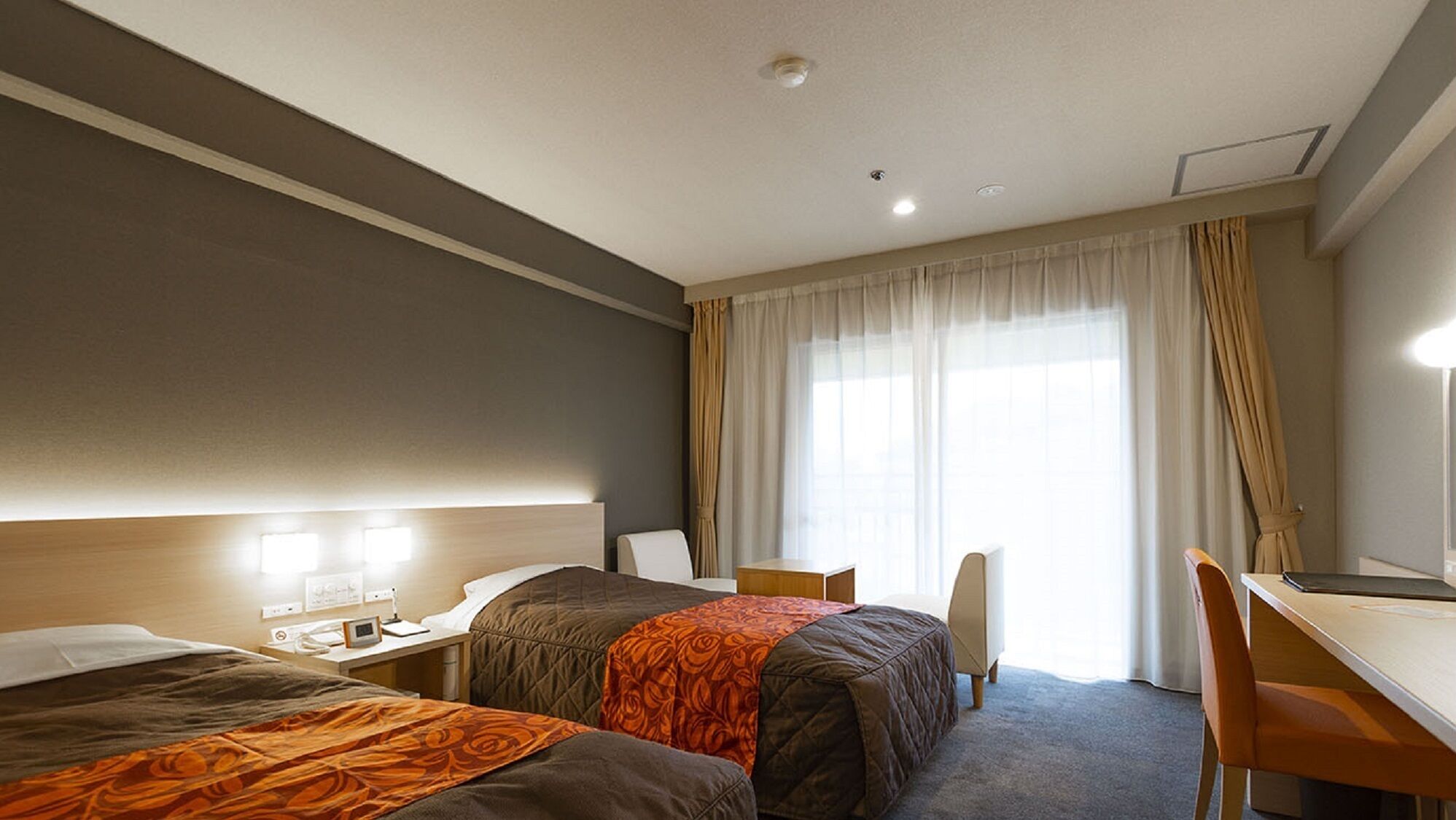 Quarto de hotel com duas camas, uma escrivaninha, uma cadeira e uma janela grande com cortinas.