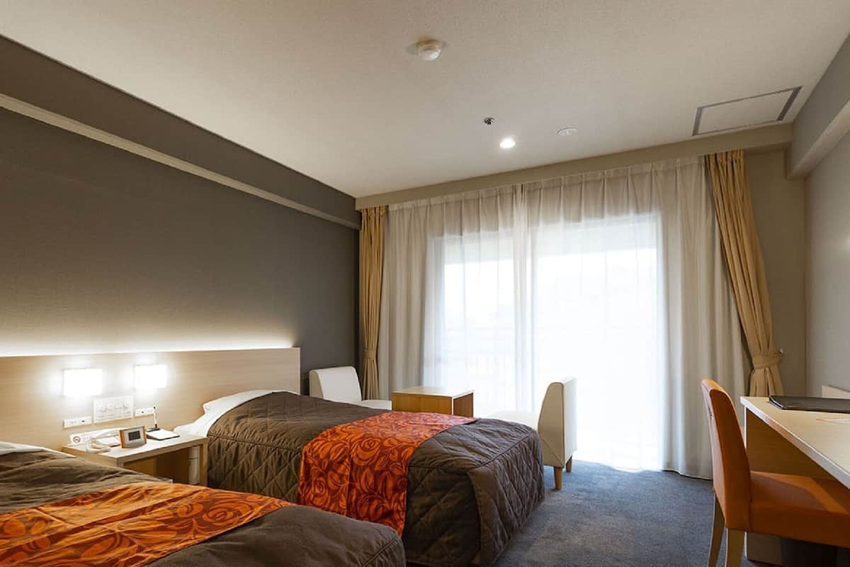 Habitación de hotel con dos camas, un escritorio, una silla y un amplio ventanal con cortinas.