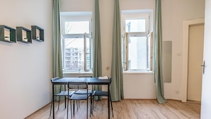 Una habitación con piso de madera, una mesa con cuatro sillas, dos ventanas con cortinas y una puerta.