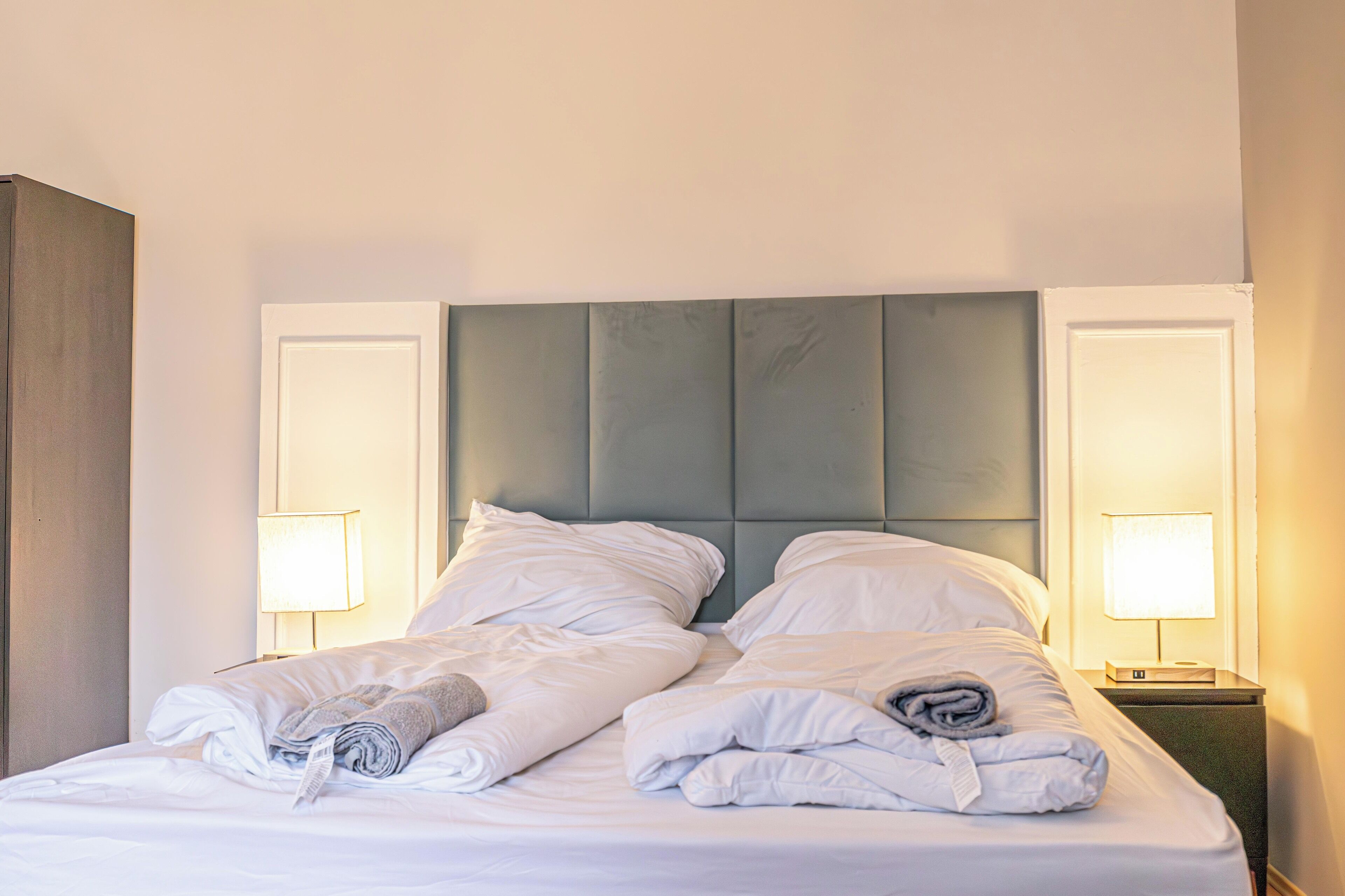 Een net opgemaakt bed met kussens en opgevouwen dekens, twee bedlampjes en een gestoffeerd hoofdbord.