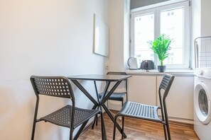 Eine kleine Küchenzeile mit Tisch und zwei Stühlen, ein Fenster mit Blick auf Gebäude, eine Topfpflanze und eine Waschmaschine.