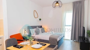 Una habitación moderna y bien iluminada con una mesa de comedor para dos personas, una cama con cabecera azul y un televisor sobre un mueble.