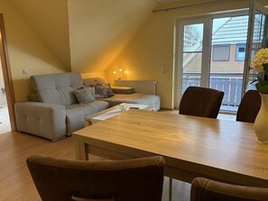 A cozy living room with a sofa, a dining table, and chairs. There is a window with a view of a house and a balcony. - Jacobihof - Strandhafer - 3-room apartment close to the beach in a quiet cul-de-sac location (Dahme)