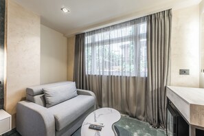 A modern hotel room with a grey sofa, a marble coffee table, and a large window with sheer curtains. - 200 meters to the main square near everything  supermarket near us 24/7 open (Tiranë)