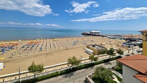 Ein Strand mit mehreren Reihen von Sonnenschirmen und Liegestühlen, eine Mole, die ins Meer führt, und ein Gebäude mit rotem Ziegeldach.