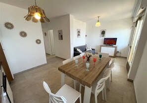 A dining area with a wooden table, white chairs, and a television on a stand. - Apartment with sea view in Lignano (Lignano Sabbiadoro)