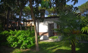 A small, single-story building with a white exterior and a door, surrounded by lush greenery and trees. - Cozy villa close to the beach (Lignano Sabbiadoro)