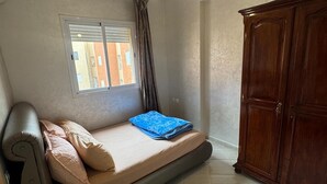 Een kleine kamer met een bed, een tafeltje, een kledingkast en een raam met zonwering.