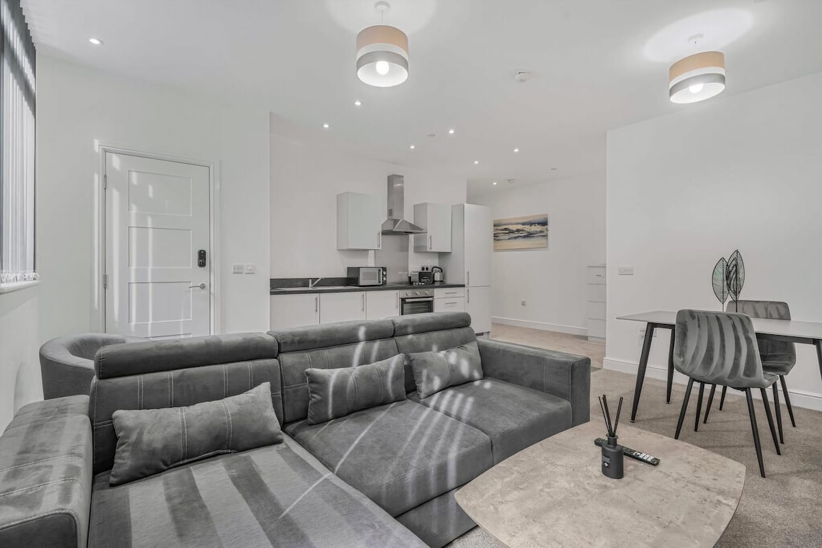 A modern living room with a grey sectional sofa, a dining table with chairs, and a kitchen in the background.