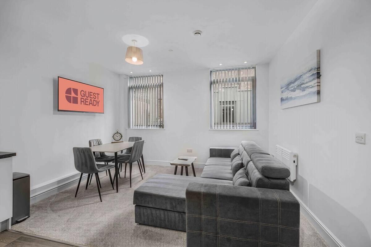 A modern living room with a dining area, grey sofas, a small table, and a 'GUEST READY' sign.
