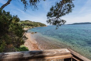 Une terrasse en bois surplombe une mer d’un bleu limpide, avec une plage de sable et une végétation luxuriante sur la côte.