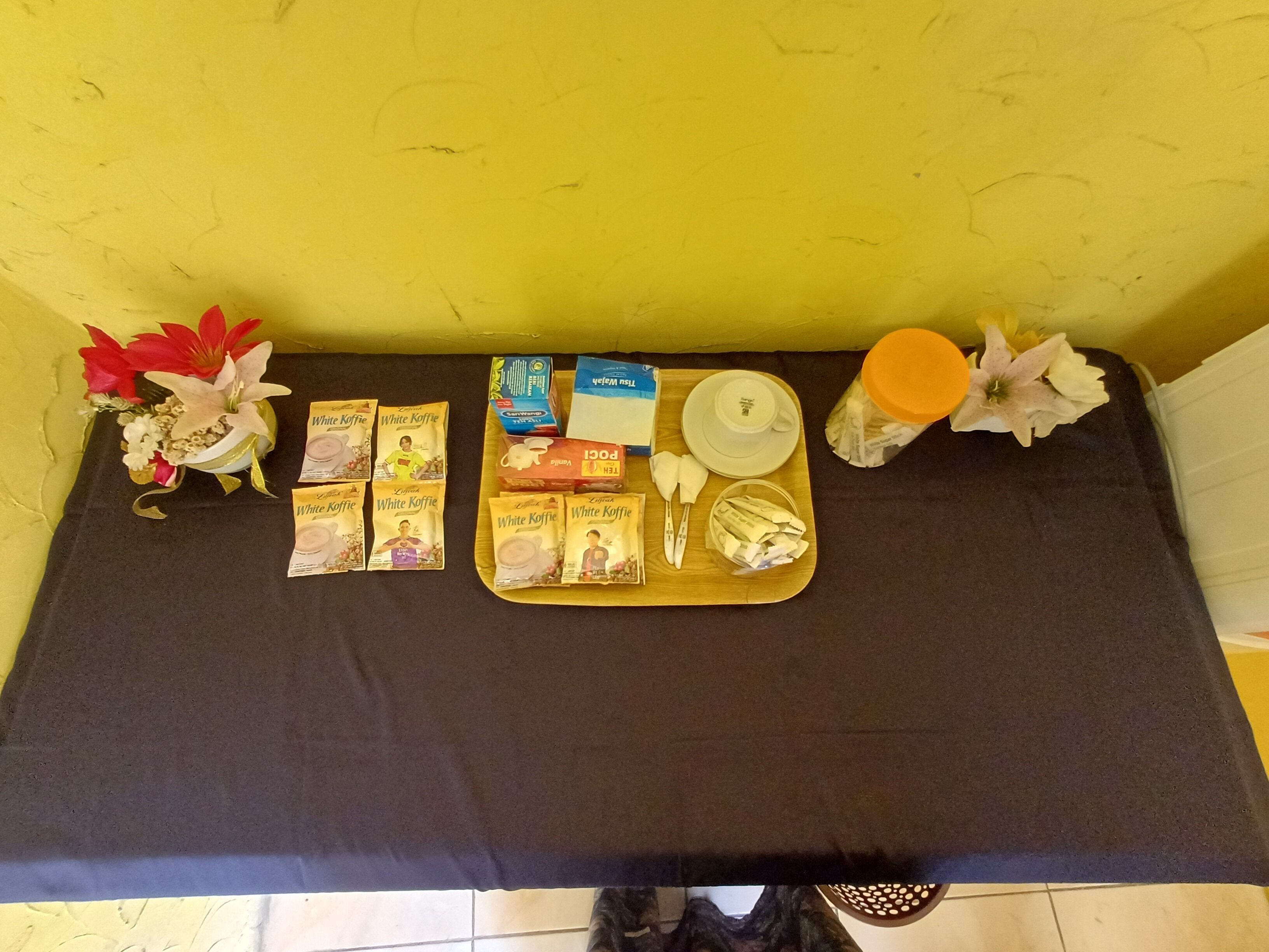 A table with a tray of white coffee packets, a box of tea, a cup, a saucer, a jar, and a vase with flowers.
