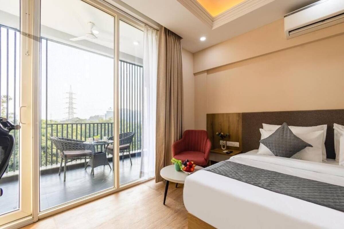 A hotel room with a large bed, a small table with fruit, a red chair, and a balcony with a view of the city.
