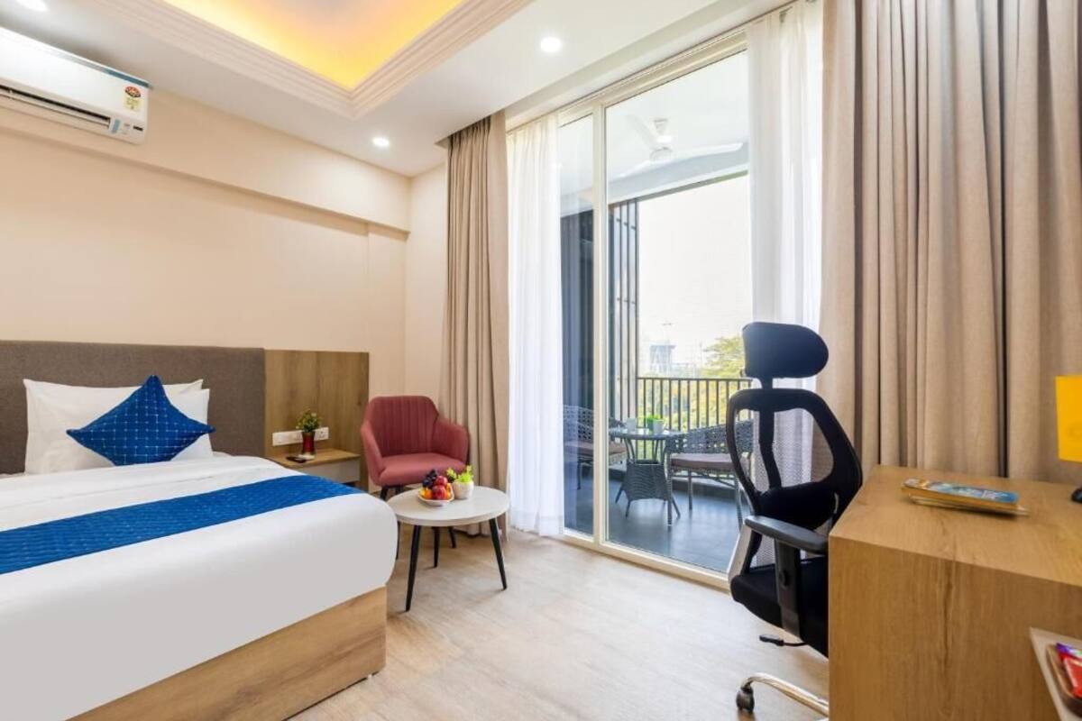 A hotel room with a bed, a desk, a chair, a small table with fruit, a red armchair, and a balcony with outdoor furniture.