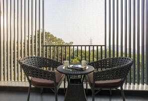 En balkon med to fletstole, et lille bord, myggenet i vinduet og udsigt til grønne omgivelser.
