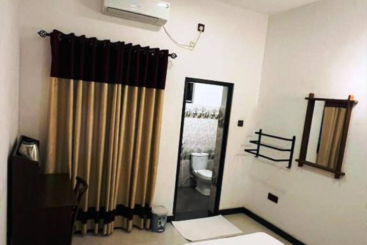 Une chambre d’hôtel comprenant un lit, un bureau, une chaise, un miroir, un ventilateur de plafond et une salle de bain visible à travers une porte ouverte.