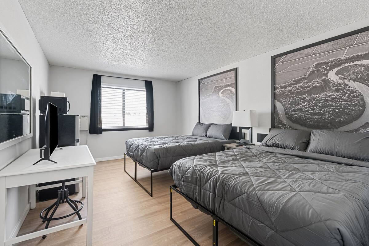 A modern bedroom with a large bed, a smaller bed, a desk with a computer, and a window with blinds.