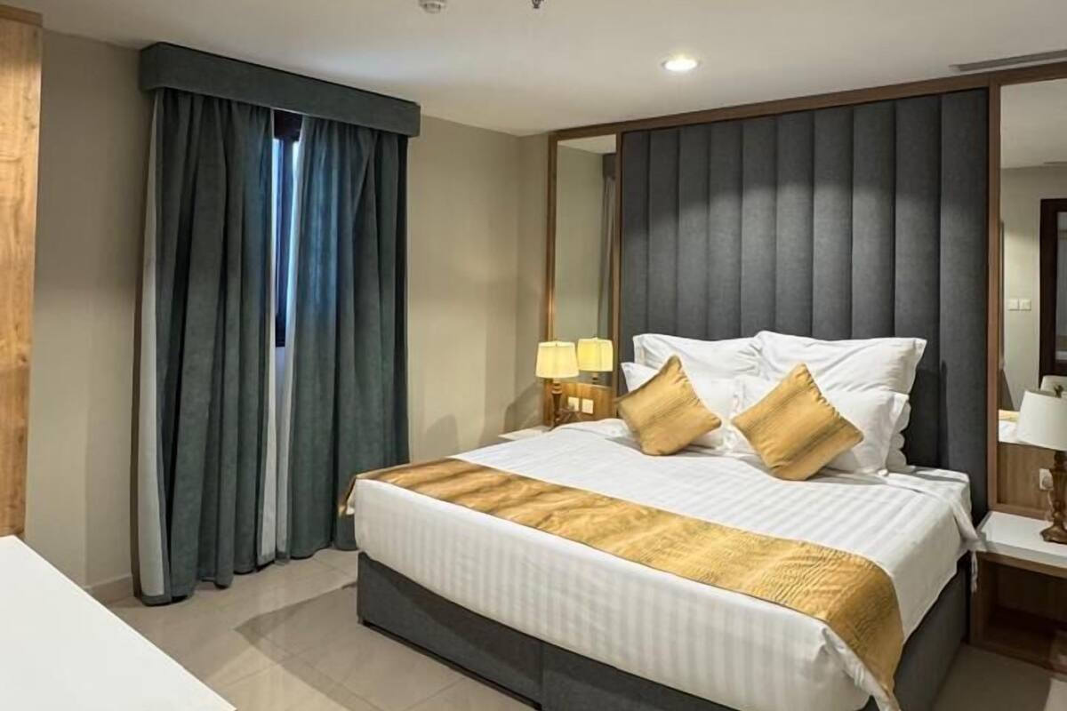 Una habitación de hotel moderna con una cama grande, una mesita de noche con lámparas, un espejo y vistas al exterior a través de la ventana.