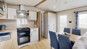 A modern kitchen with a stove, oven, and cabinets, a dining area with a table and chairs, and a window with curtains. - Gold 3 Bedroom Holiday Home (Hornsea)
