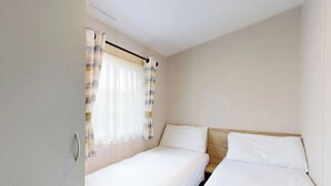 Una habitación pequeña con cama individual, cabecera de madera, ventana con cortinas de gasa y una puerta a la izquierda.