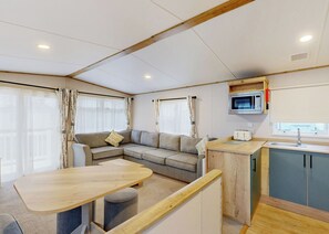 A compact living space with a sofa, a small table, a kitchenette with a microwave, and a window with curtains. - Silver 3 Bedroom Holiday Home Pet (Hornsea)